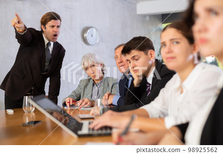 Displeased male manager scolding at young, adult, mature office workers sitting with reports and laptops at table in meeting room 98925562