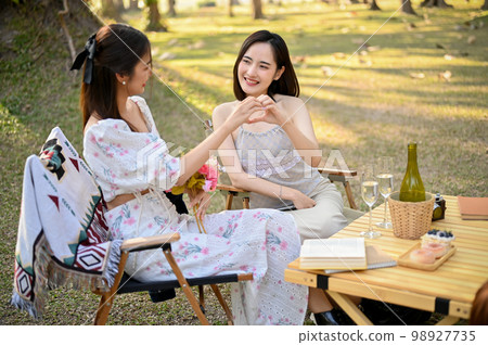 Attractive Asian woman making a heart hand sign with her friend while picnicking in the park 98927735