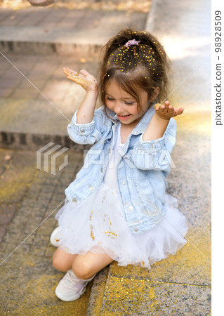 Funny girl strewn with small golden confetti sequins. The child is sitting in the park on the steps laughing merrily 98928509