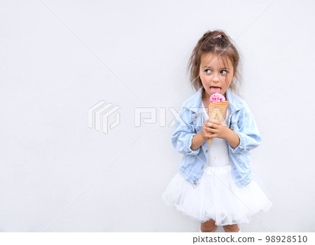 A funny girl stands at the city white wall and licks a pink ball of ice cream looking away. Banner with copy space A funny girl stands at the city white wall and licks a pink ball of ice cream looking away. Banner with copy space 98928510