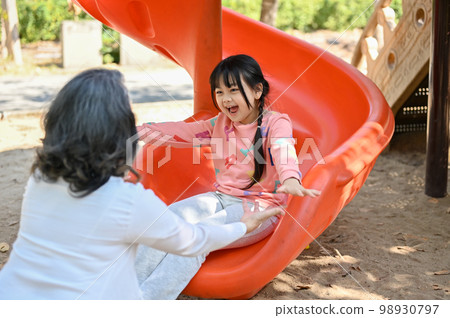 Happy Asian little girl playing on slider, enjoys playing on playground with her grandmother Happy Asian little girl playing on slider, enjoys playing on playground with her grandmother 98930797