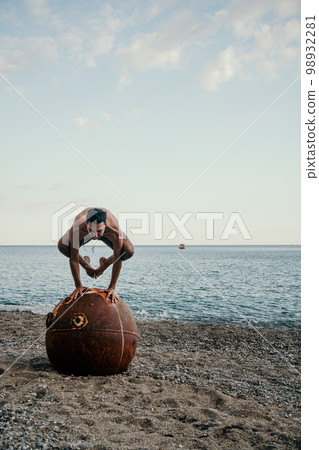Man doing yoga exercise outdoors standing on old rusty floating marine mine on the beach with rocky shore and sea background. Healthy lifestyle, pollution, nature protection, war and peace concept. 98932281
