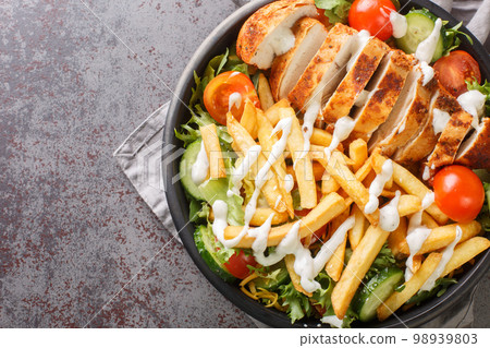 Pittsburgh steak salad consist a lettuce salad with cheese, chicken breast, French fries, and ranch dressing on top closeup on the bowl. Horizontal top view Pittsburgh steak salad consist a lettuce salad with cheese, chicken breast, French fries, and ranch dressing on top closeup on the bowl. Horizontal top view 98939803