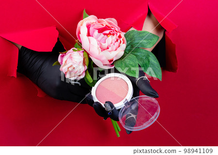 Gloved hands with round powder box and flowers. Breaking through a red paper wall. Gloved hands with round powder box and flowers. Breaking through a red paper wall. 98941109