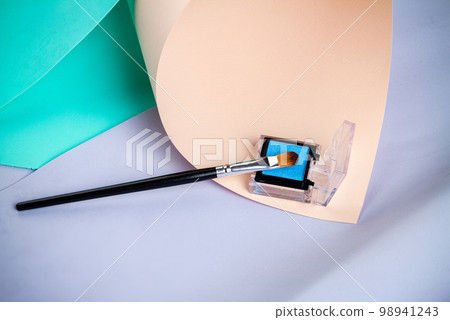Square shape cosmetic box with blue powder. Male up brush with black handle. Colored papers background. Square shape cosmetic box with blue powder. Male up brush with black handle. Colored papers background. 98941243