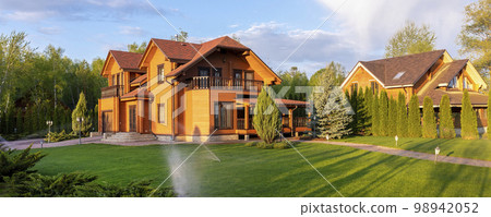 Modern wooden eco house villa facade luxury big house. Timber cottage with with green lawn water sprinkler, paved footpath and blue sky background. Landscaping design, garden watering and maintenance 98942052