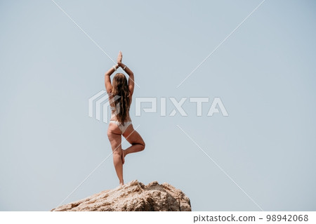 Woman sea yoga. Back view of free calm bliss satisfied woman with long hair standing on top rock with yoga position against of sky by the sea. Healthy lifestyle outdoors in nature, fitness concept. Woman sea yoga. Back view of free calm bliss satisfied woman with long hair standing on top rock with yoga position against of sky by the sea. Healthy lifestyle outdoors in nature, fitness concept. 98942068