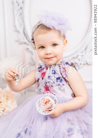 Baby girl elegant dress. A one-year-old girl in a puffy dress and a cute bow poses against the backdrop of a bright room with a dressing table and flowers. 98943922
