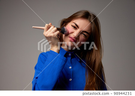 Half-length portrait of young emotional beautiful girl holding blush brush and doing makeup isolated over grey background. Beauty, fashion, style, fun and joy concept Half-length portrait of young emotional beautiful girl holding blush brush and doing makeup isolated over grey background. Beauty, fashion, style, fun and joy concept 98944231