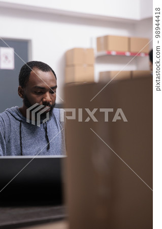 Storehouse manager checking shipment logistics before start preparing packages, putting customer order in carton boxes. African american supervisor working in warehouse delivery department Storehouse manager checking shipment logistics before start preparing packages, putting customer order in carton boxes. African american supervisor working in warehouse delivery department 98944418