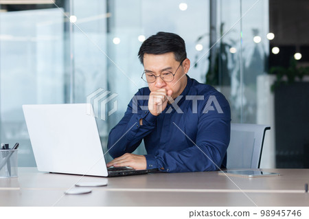 Asian businessman sick working at workplace inside office with laptop, man in shirt and glasses coughs and has a cold. 98945746