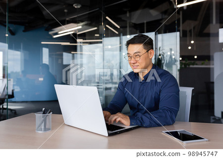 Successful mature asian working inside office using laptop, man typing on keyboard and smiling, businessman in shirt and glasses satisfied with work and achievement results, programmer at work. 98945747