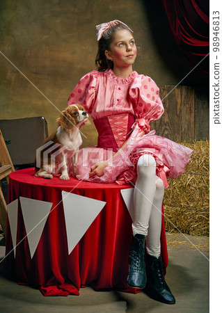 Dreaming. Beautiful little girl in festive pink dress posing with cute dog over dark vintage circus background. Holidays, dreams, art, fashion, vintage style 98946813