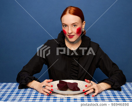 Beetroot cheeks. Young beautiful girl with beet on retro tablecloth over blue studio background. Food pop art photography. Complementary colors 98946898