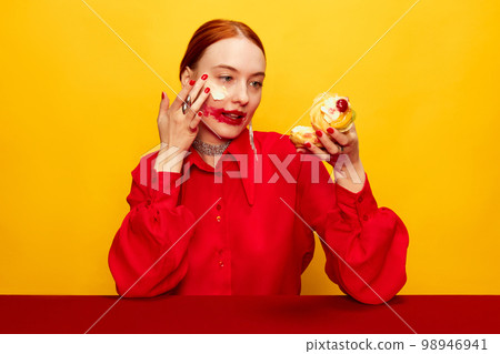 Surrealism. Beautiful young girl in red shirt with smudged red lipstick, eating cake over yellow studio background. Food pop art photography. Complementary colors 98946941