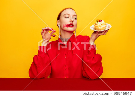 Beautiful young girl in red shirt tasting cake with cherry over yellow studio background. Food pop art photography. Complementary colors Beautiful young girl in red shirt tasting cake with cherry over yellow studio background. Food pop art photography. Complementary colors 98946947