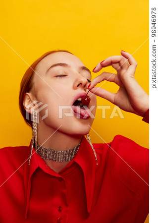 Beautiful young girl in red shirt eating sweet cherry from dessert over yellow studio background. Food pop art photography. Complementary colors Beautiful young girl in red shirt eating sweet cherry from dessert over yellow studio background. Food pop art photography. Complementary colors 98946978