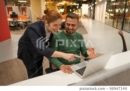 A red-haired Caucasian woman stands at the desktop of a bearded man. The boss approves the work of the subordinate. A red-haired Caucasian woman stands at the desktop of a bearded man. The boss approves the work of the subordinate. 98947149
