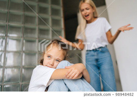 Mad blonde abusive mother emotionally gesturing and shouting at crying little child daughter kid sitting hugging knee, depressed looking away. Concept of scandal, crisis in family, domestic violence. Mad blonde abusive mother emotionally gesturing and shouting at crying little child daughter kid sitting hugging knee, depressed looking away. Concept of scandal, crisis in family, domestic violence. 98948482