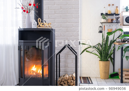 Valentine decor near fireplace stove with fire and firewood. Cozy home hearth in interior with potted plans, valentines day in family love nest. Valentine decor near fireplace stove with fire and firewood. Cozy home hearth in interior with potted plans, valentines day in family love nest. 98949036