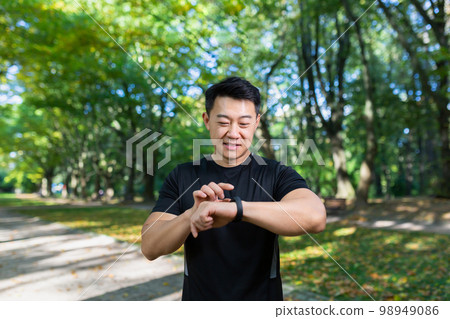 Smiling and successful asian man jogging and exercising in the park, man looking at smart watch and comparing results, athlete happy with results and smiling. 98949086