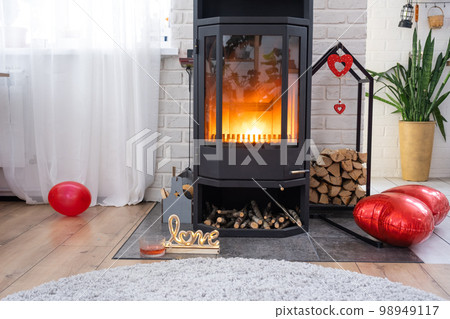 Valentine decor near fireplace stove with fire and firewood. Cozy home hearth in interior with potted plans, valentines day in family love nest. Valentine decor near fireplace stove with fire and firewood. Cozy home hearth in interior with potted plans, valentines day in family love nest. 98949117