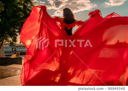 Sunrise red dress. A woman in a long red dress against the backdrop of sunrise, bright golden light of the sun's rays. The concept of femininity, harmony. 98949348