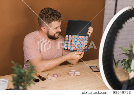 Man talking on cosmetics holding a makeup tools while recording his video. Guy making video for his blog on cosmetic product Man talking on cosmetics holding a makeup tools while recording his video. Guy making video for his blog on cosmetic product 98949798