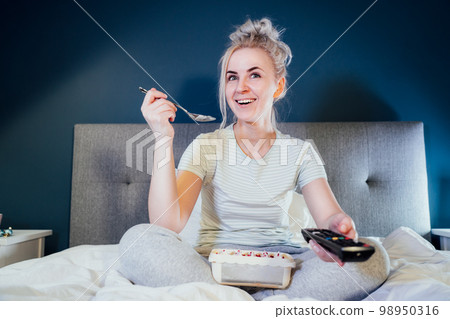 Young cheerful woman in home clothes eating cake and watching movie on cable TV while switching channels with the remote control in her bed at home in evening alone. Leasure, relaxation time Young cheerful woman in home clothes eating cake and watching movie on cable TV while switching channels with the remote control in her bed at home in evening alone. Leasure, relaxation time 98950316