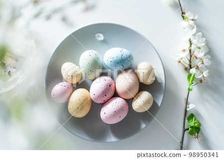 Top view colored easter eggs on the gray plate, blooming tree branch in vase and flower petals on the white table. Happy Easter and springtime card. Minimalism and simplicity. Selective focus. 98950341