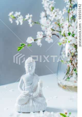 Decorative white Buddha statuette with blooming tree branches in the vase on the light background. Meditation and relaxation ritual. Buddha birthday. Vertical card. Selective focus Decorative white Buddha statuette with blooming tree branches in the vase on the light background. Meditation and relaxation ritual. Buddha birthday. Vertical card. Selective focus 98950342