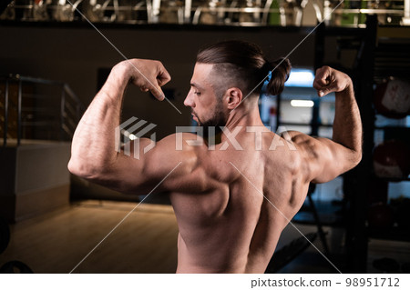 A handsome bearded male athlete with a muscular body poses and shows his biceps and back in the gym A handsome bearded male athlete with a muscular body poses and shows his biceps and back in the gym 98951712