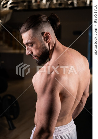 A handsome bearded male athlete with a muscular body poses in the gym in white pants. Active lifestyle, sports A handsome bearded male athlete with a muscular body poses in the gym in white pants. Active lifestyle, sports 98951716