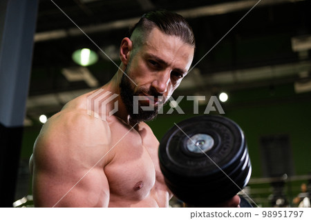 A brutal muscular bearded man holds a dumbbell in the gym and shows his biceps. Active lifestyle, sports A brutal muscular bearded man holds a dumbbell in the gym and shows his biceps. Active lifestyle, sports 98951797