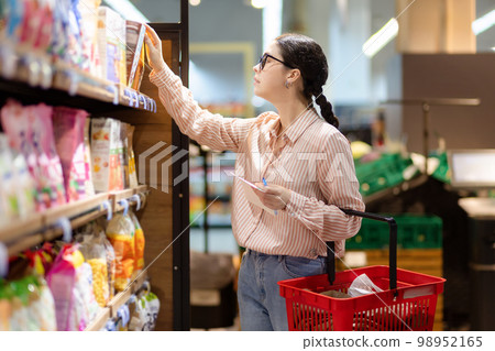 Side view of young attractive caucasian woman wearing eyeglasses holds cart and reach hand to take food from top shelf. Concept of shopping in supermarket and consumerism 98952165