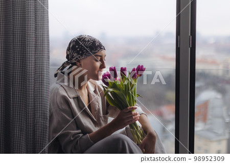 Young woman with cancer sitting in a window and smelling flowers. 98952309