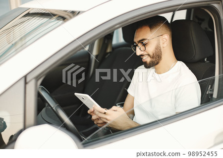 Handsome indian young man looking at his smart phone while sitting in the car. Man wearing white t-shirt and glasses. Male driver sitting on car and hand holding smartphone to use online application. Handsome indian young man looking at his smart phone while sitting in the car. Man wearing white t-shirt and glasses. Male driver sitting on car and hand holding smartphone to use online application. 98952455