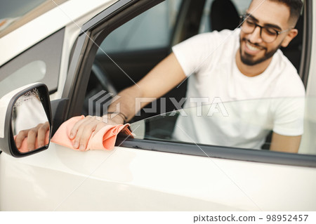 Young man in glasses polishing inside his car with a rug. Man wearing white t-shirt. Indian man wiping his white vehicle. Young man in glasses polishing inside his car with a rug. Man wearing white t-shirt. Indian man wiping his white vehicle. 98952457