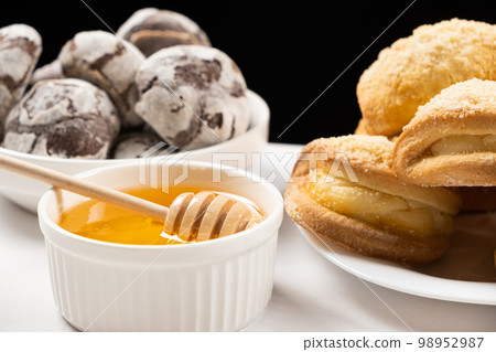 Liquid natural bee honey in a white ceramic bowl with a wooden spoon, with honey cookies and cocoa cookies. 98952987