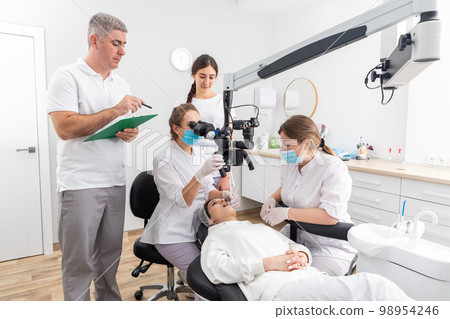 Group of young doctors learning in dental clinic watching dental treatment with microscope Group of young doctors learning in dental clinic watching dental treatment with microscope 98954246
