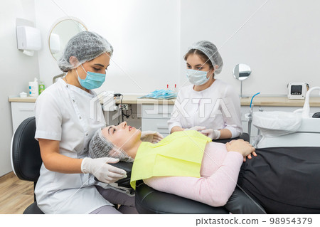 Dentist and assistant performing dental treatment inmodern dental clinic, patient laying in a chair 98954379