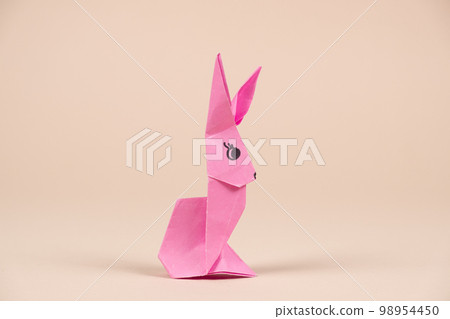 A pink origami rabbit on a beige background. Crafts for Easter, fold from paper, do it yourself. A pink origami rabbit on a beige background. Crafts for Easter, fold from paper, do it yourself. 98954450