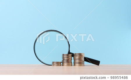 A stack of metal coins and a plastic magnifier on a blue background. The concept of increasing taxes, subsidies 98955486