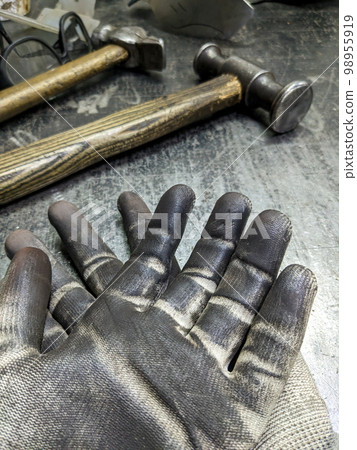 Black blacksmith work gloves against a sledgehammer background. Hard men's work as a blacksmith. 98955919