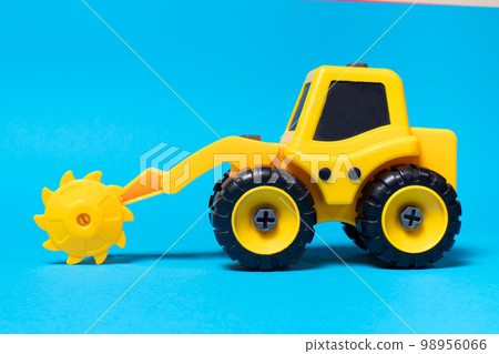 Toy yellow tractor with a round saw nozzle for wood on a blue background. Construction machine appliances for the toy store Toy yellow tractor with a round saw nozzle for wood on a blue background. Construction machine appliances for the toy store 98956066