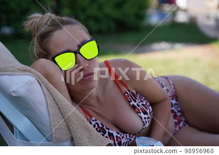 A curvy-breasted blonde in a colorful swimsuit and chroma key sunglasses lies on her side on the beach and stares at you intently. Portrait of a young woman on the beach. 98956968