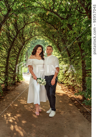 Portrait of the happy bride and groom under the green flowering arch close-up. A joyful couple in the spring garden, a girl with a bouquet of white lush roses. Portrait of the happy bride and groom under the green flowering arch close-up. A joyful couple in the spring garden, a girl with a bouquet of white lush roses. 98957048