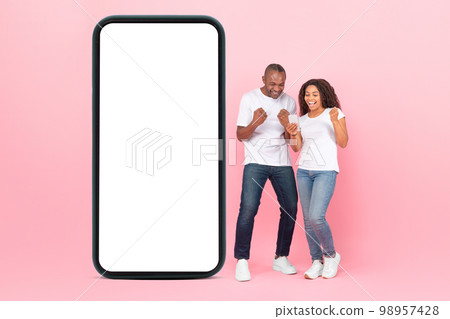 Excited african american spouses standing near huge smartphone with empty screen, using phone and shaking clenched fists Excited african american spouses standing near huge smartphone with empty screen, using phone and shaking clenched fists 98957428