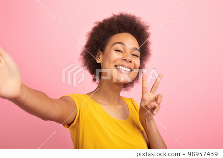 Glad excited millennial multiethnic woman in casual taking selfie and show peace sign with hand Glad excited millennial multiethnic woman in casual taking selfie and show peace sign with hand 98957439