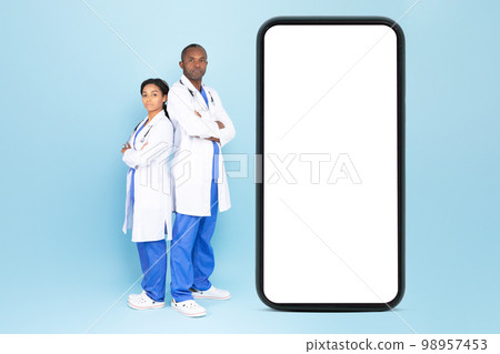 Black male and female doctors in white coats posing near big smartphone with blank screen, blue background, mockup Black male and female doctors in white coats posing near big smartphone with blank screen, blue background, mockup 98957453